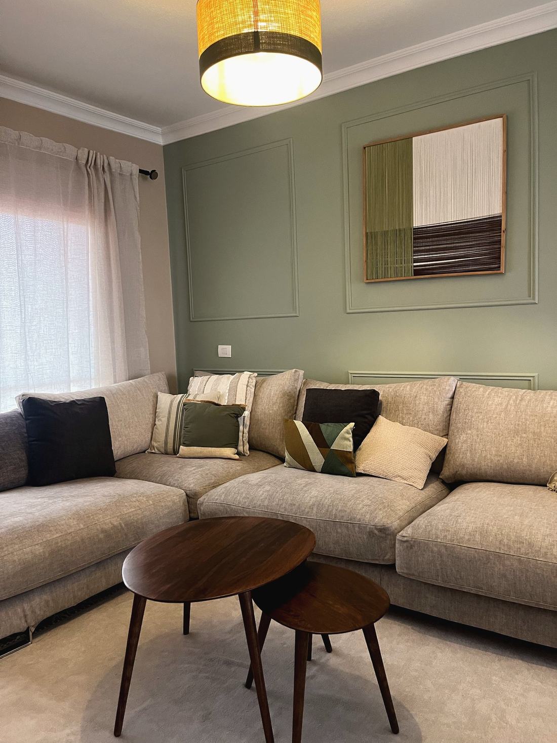 Salón con sofá en L, cuadro tejido en tonos verdes y mesas nido de madera